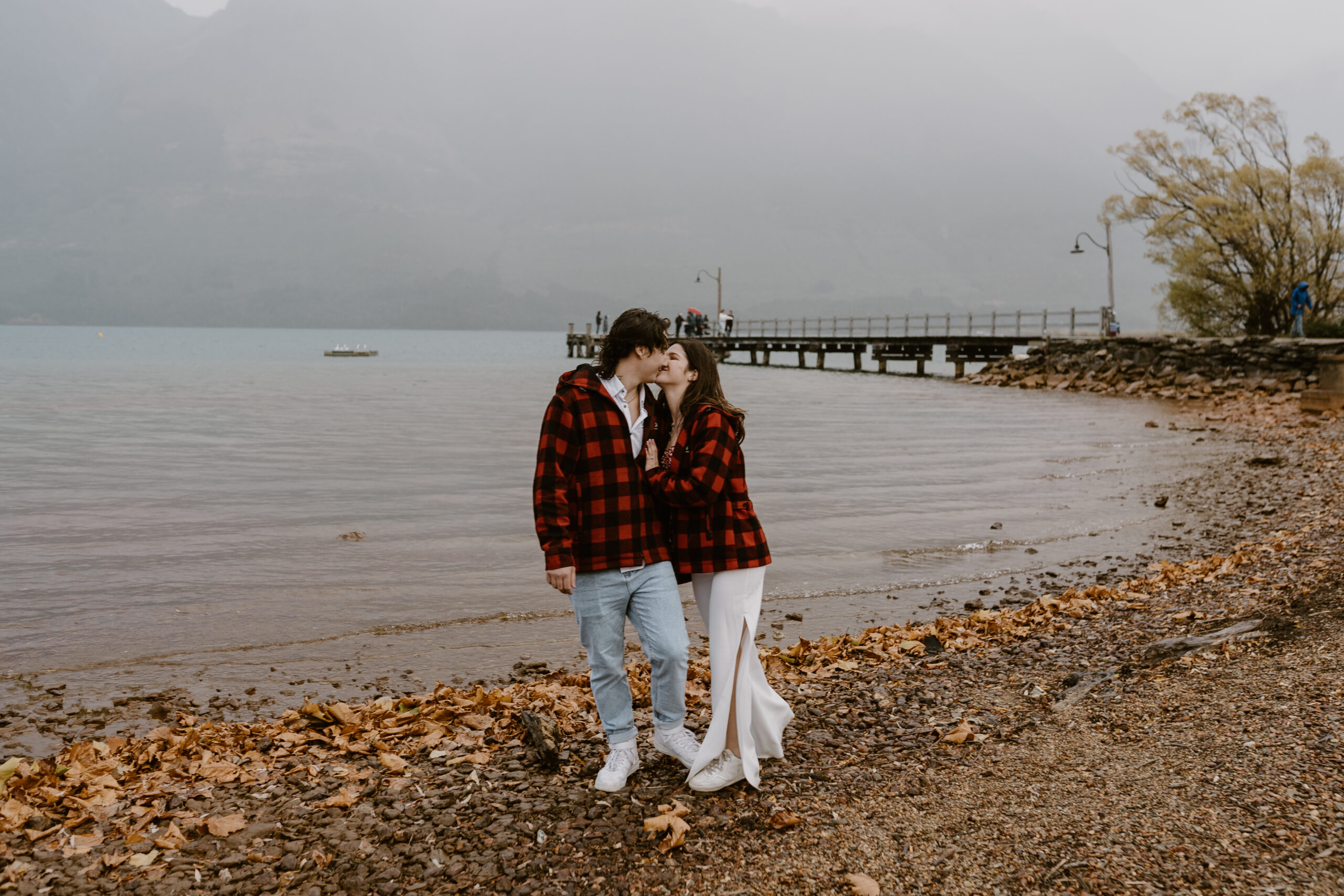 Engaged couple stop and kiss for engagement photo on lake edge in Glenorchy